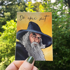 An illustrated portrait of Gandalf on a sticker that says Do Epic Shit. The sticker is held up against a leafy background that suggests The Shire, but is really the backyard at the illustrator's former home. Ooh, trivia!