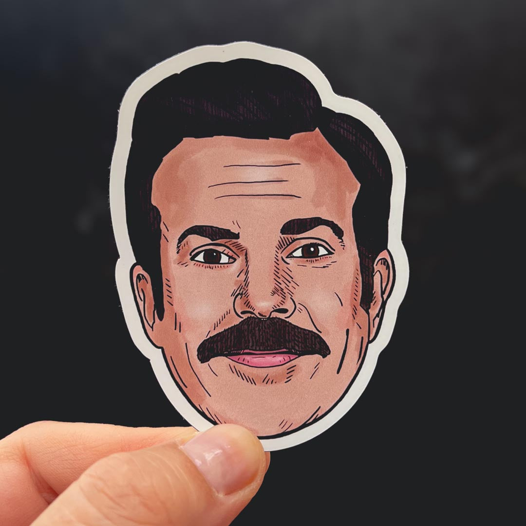 A sticker depicting Ted Lasso's smiling face held against a black backdrop