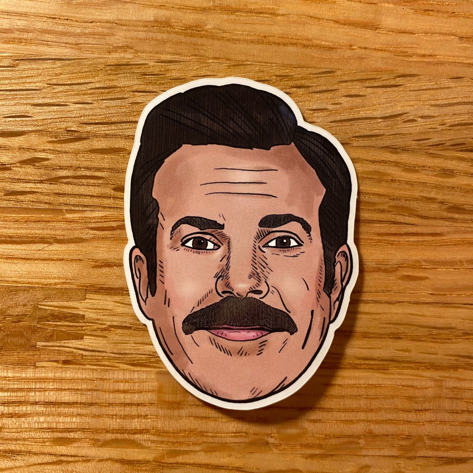 A sticker of Ted Lasso's smiling face sits on a light wood table